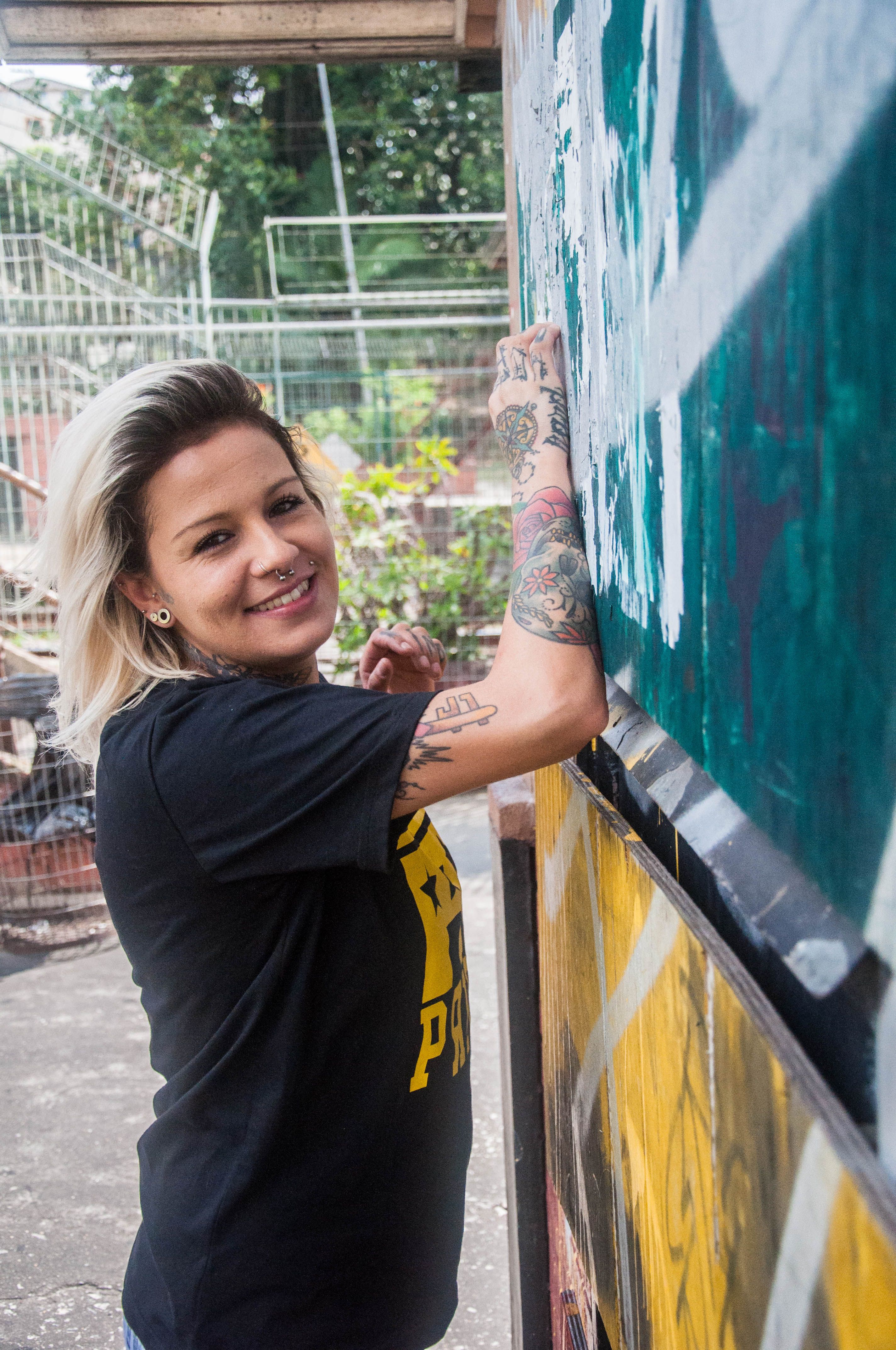 Smiling blonde woman with tattoos painting a wall outdoors.