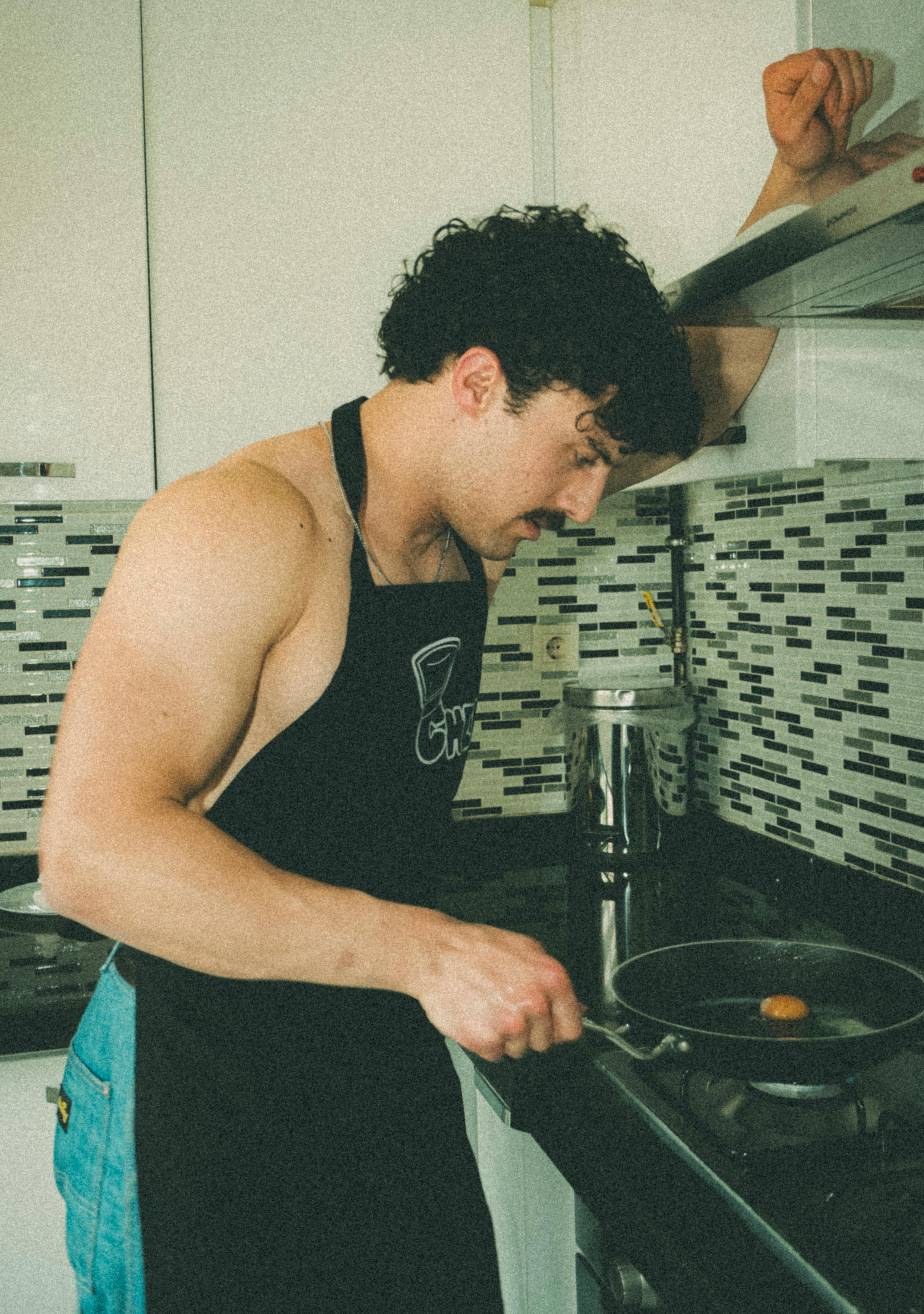 Man wearing apron cooks an egg in a modern Ankara kitchen. Styled in a warm portrait with mosaic tiles.