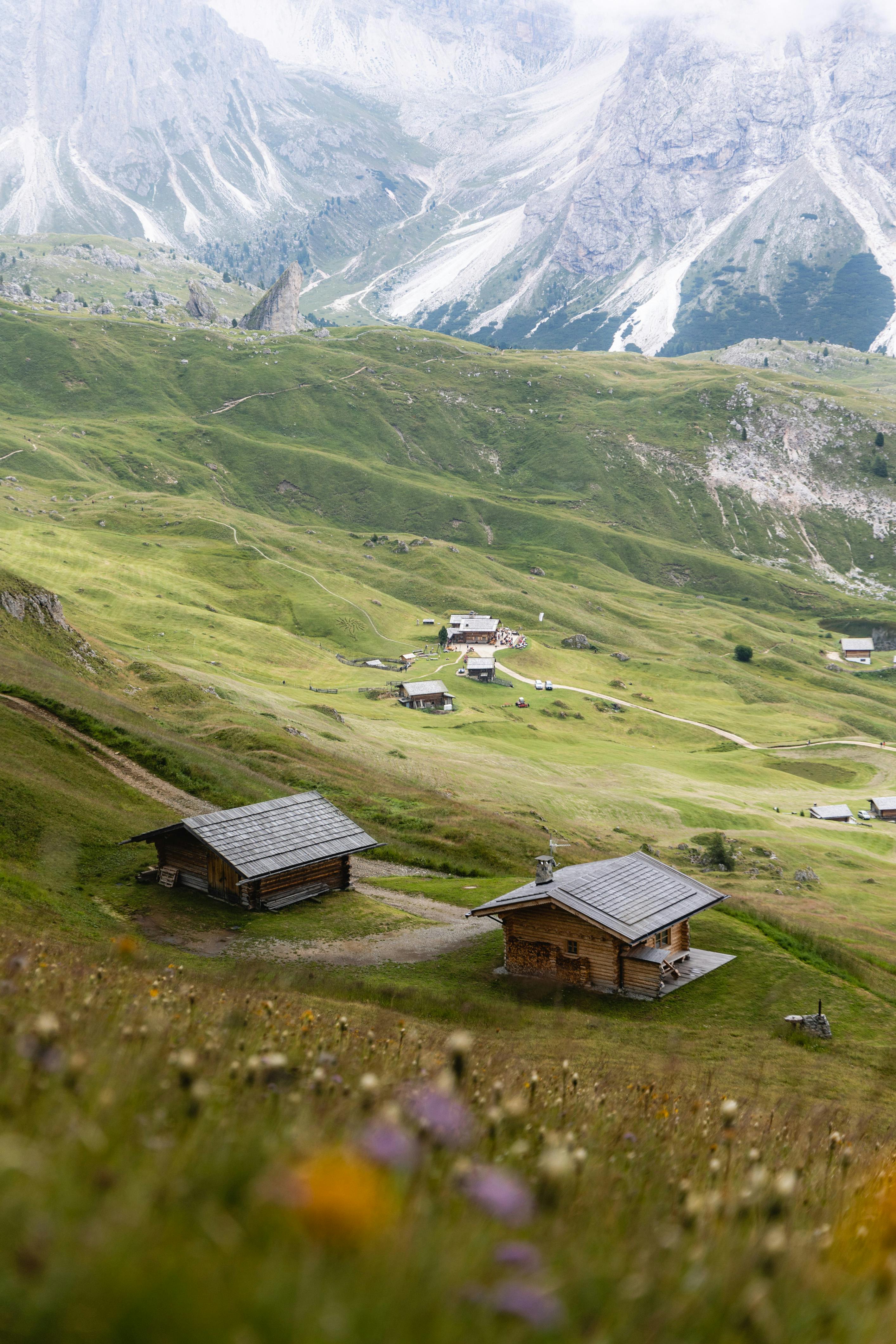 Charming wooden cabins nestled in a peaceful mountain valley with grassy hills and distant peaks.