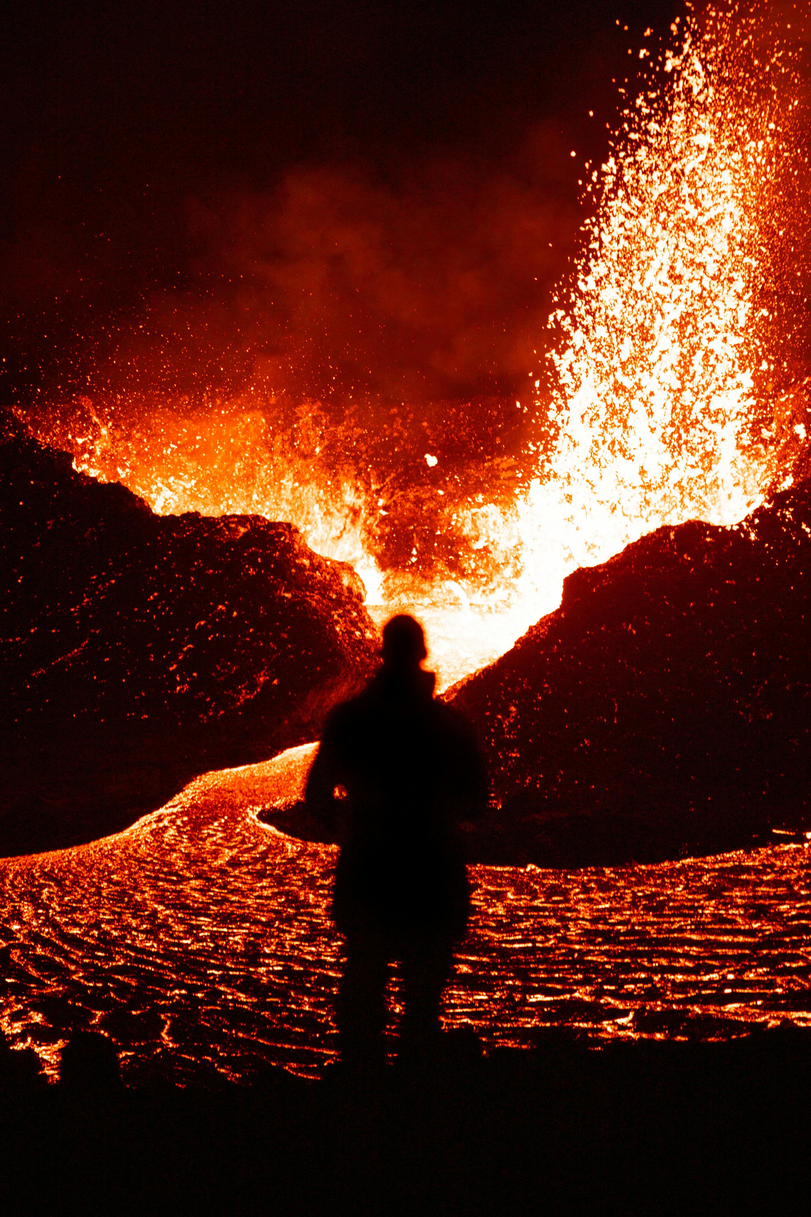 A silhouette stands before a vibrant eruption, showcasing glowing lava and intense flames.