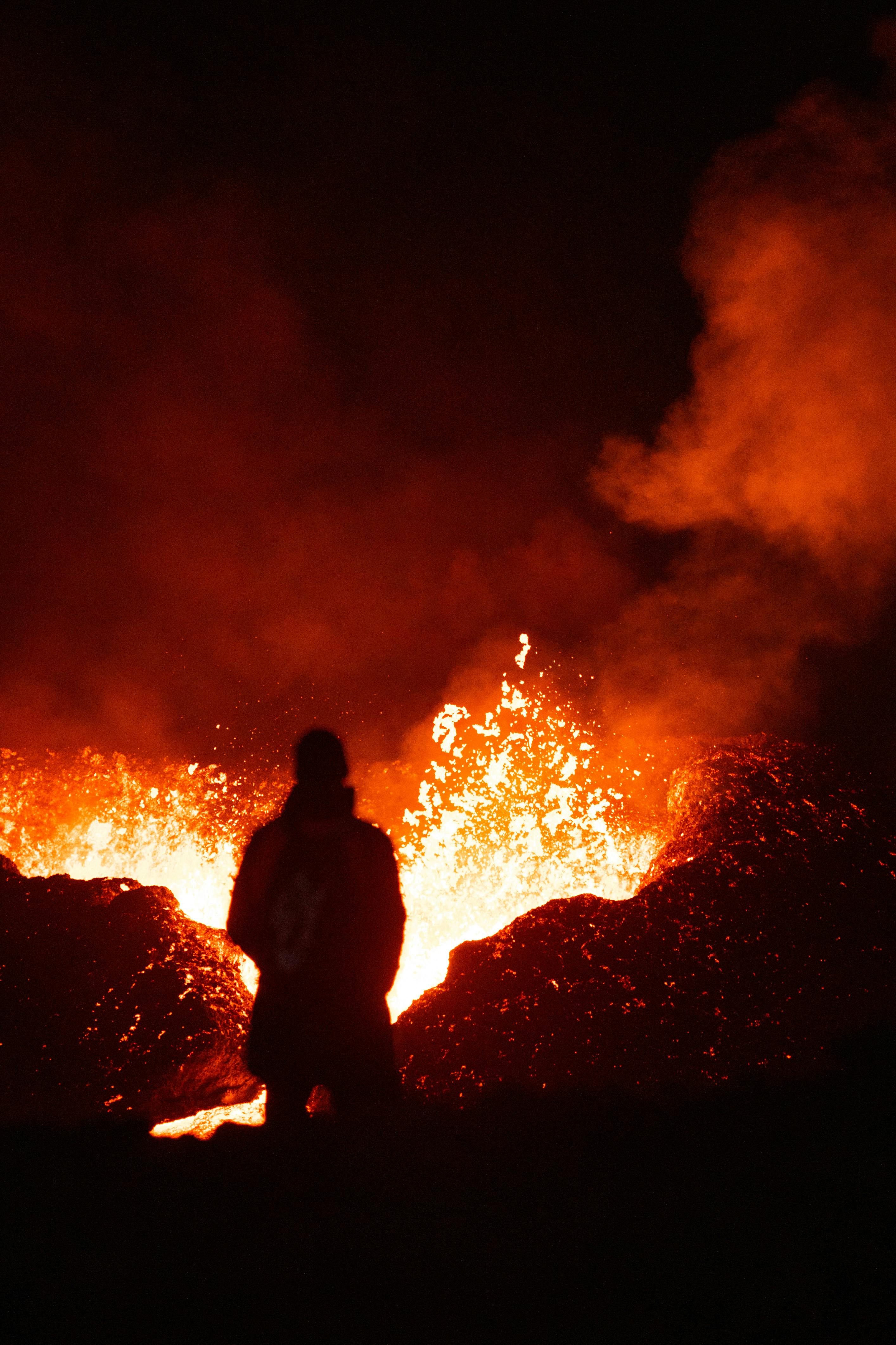 Stunning silhouette of a person against the backdrop of a volcanic eruption at night, highlighting fiery lava and smoke.