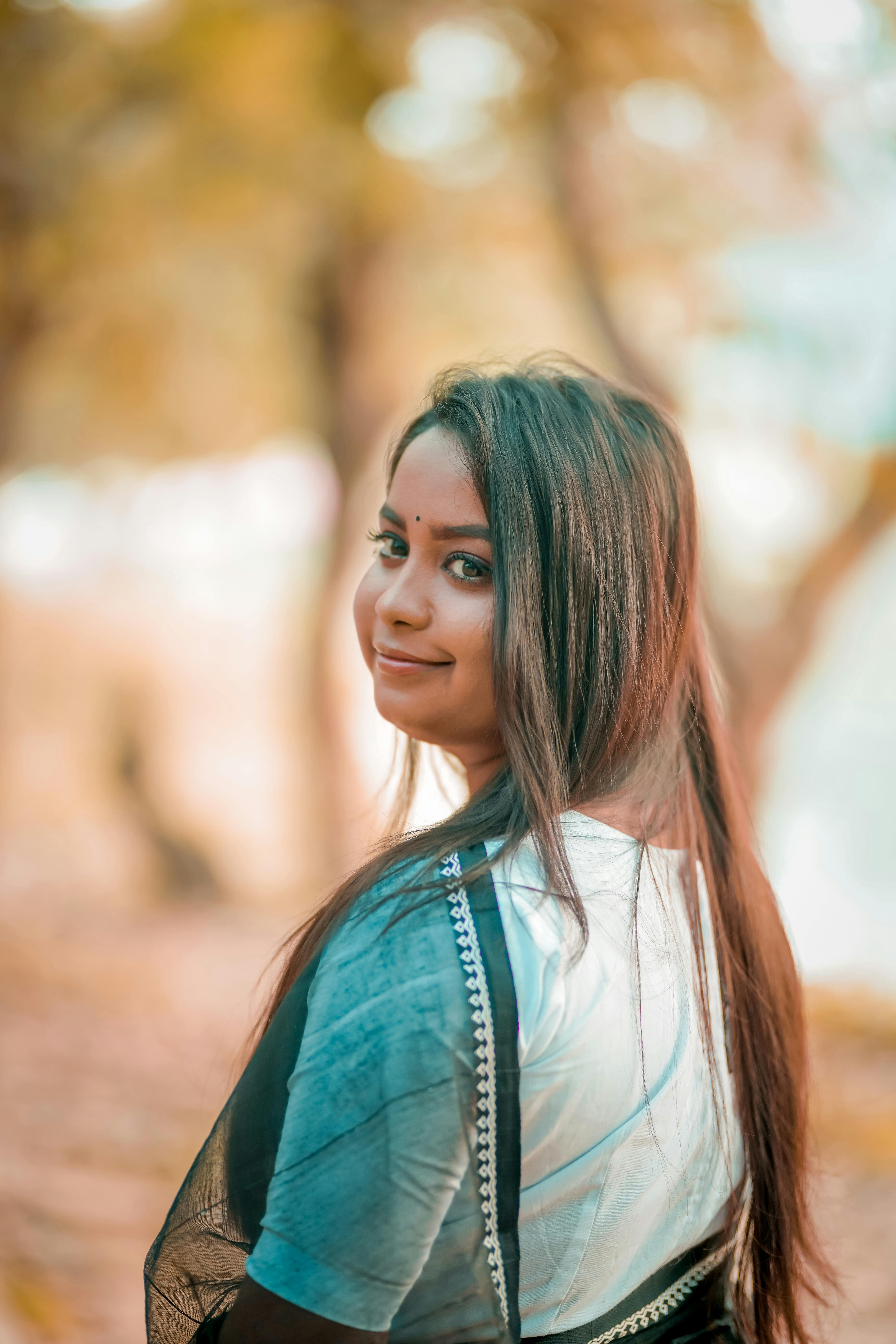 Portrait of a smiling woman with long brunette hair wearing traditional attire outdoors in a natural setting.