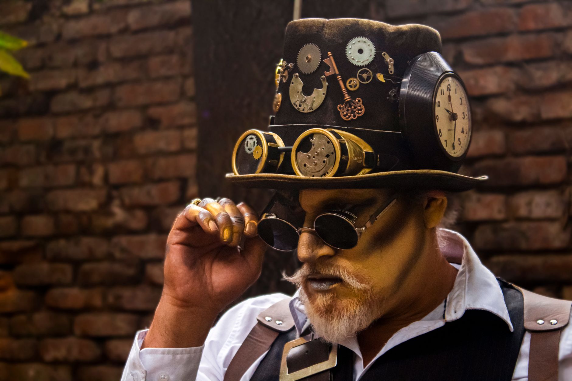 Man in steampunk attire with gears and clock hat against a brick wall background.
