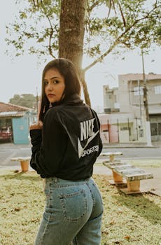 白天，一位穿休闲装的年轻女子在城市公园的树旁摆出户外姿势