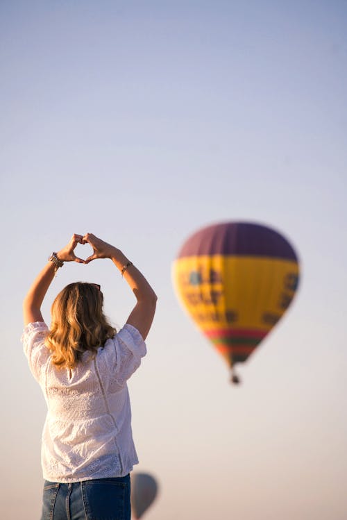 A woman making a heart shape with her hands while looking at a hot air balloon