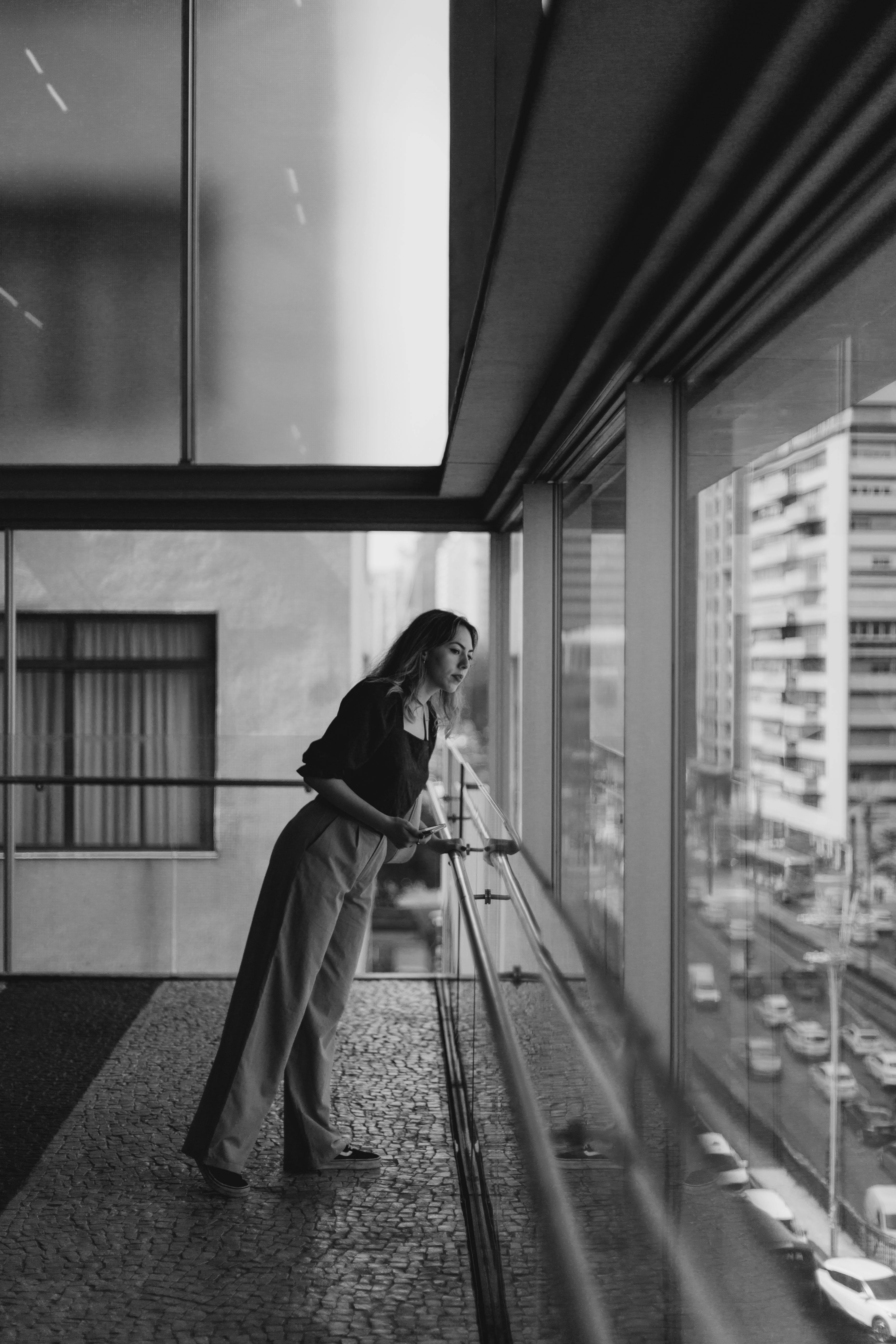 Black and white photo of a woman standing indoors by a window, overlooking urban São Paulo.