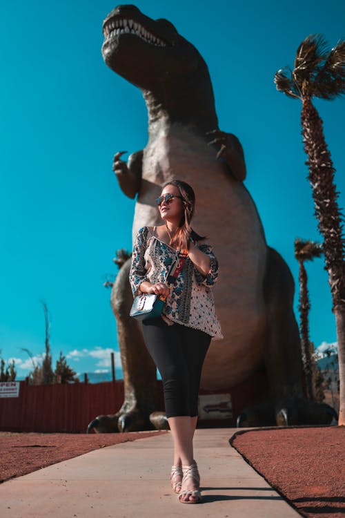 Photo of a woman wearing a printed top and sunglasses standing  near dinosaur figurine