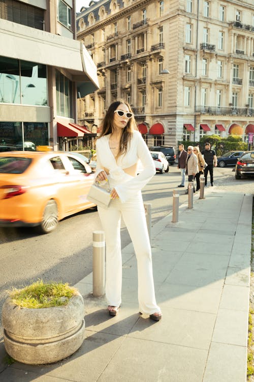A woman in white pants and a white top is standing on the sidewalk