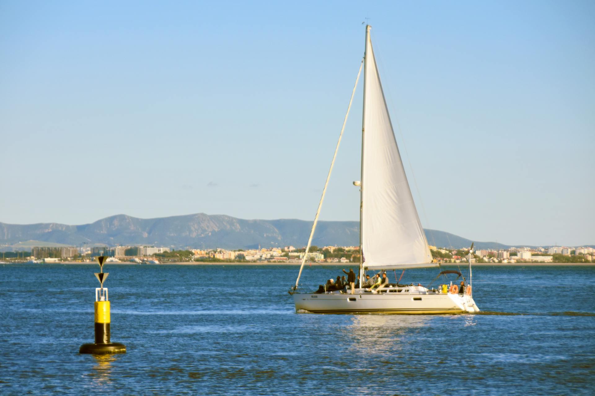 A sailboat cruises Lisbon waters under a clear sky, with mountains in the background.