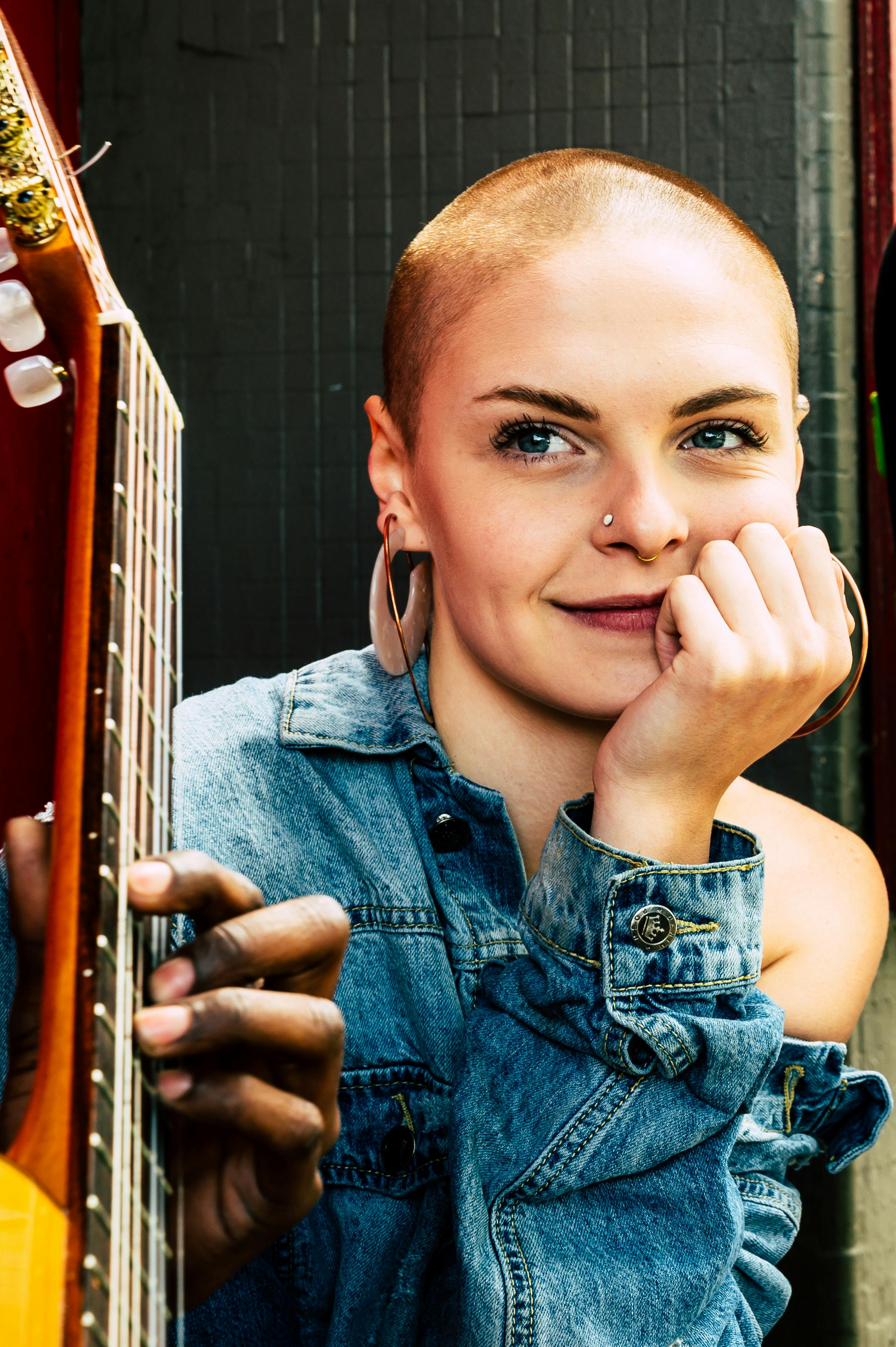 A stylish woman with a shaved head posing with a guitar, wearing a nose ring and earrings.