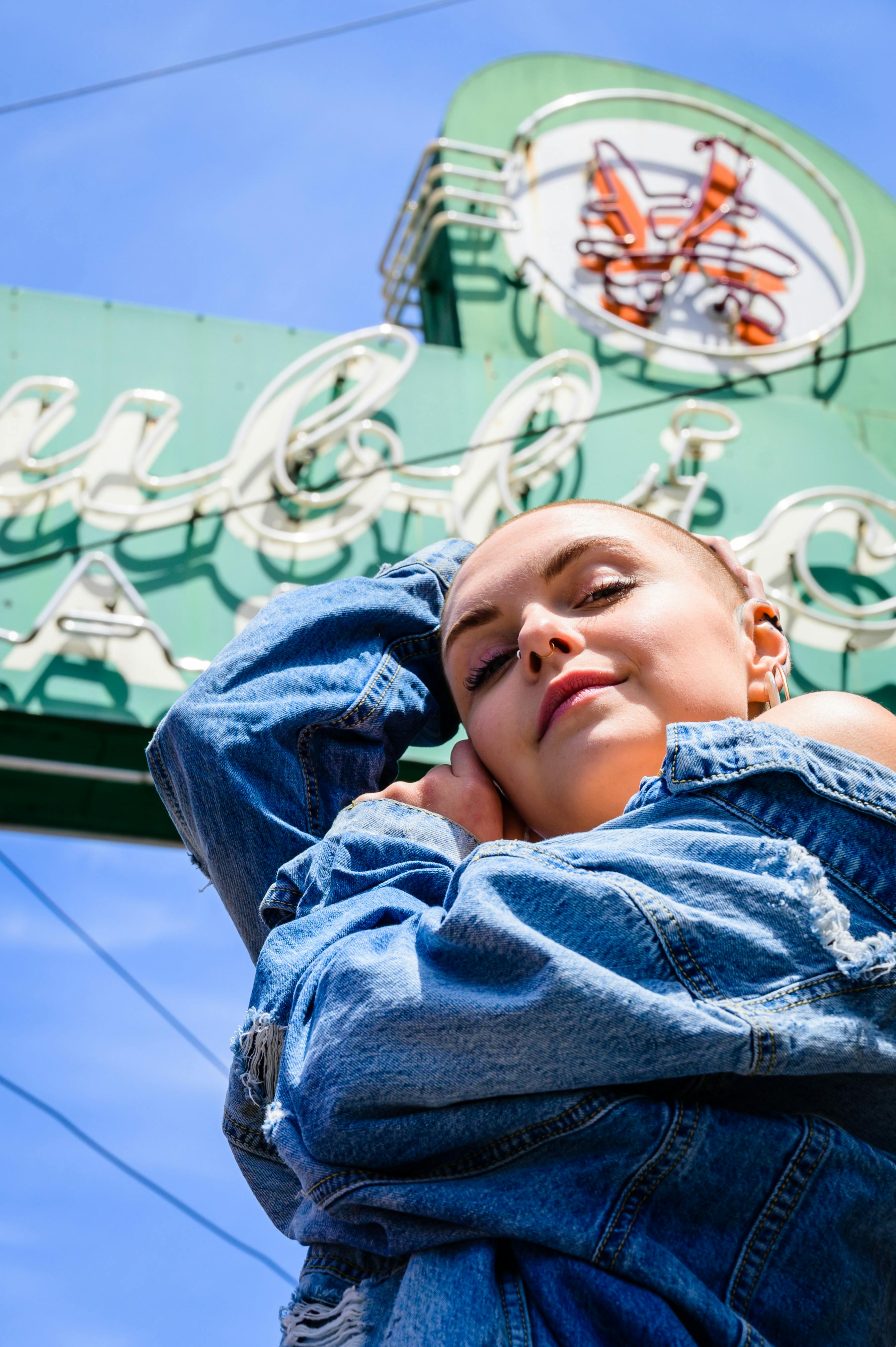 Young woman in denim posing outdoors under a vintage neon sign with a clear blue sky.