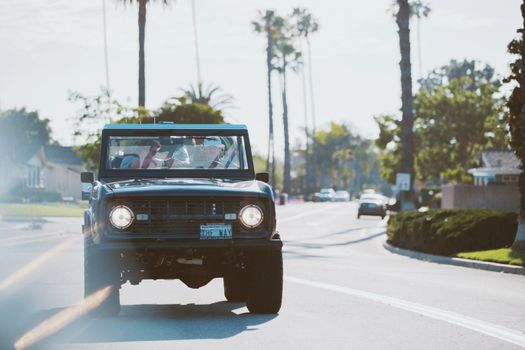 A vintage SUV cruising down a sunlit street with palm trees and houses in the background.