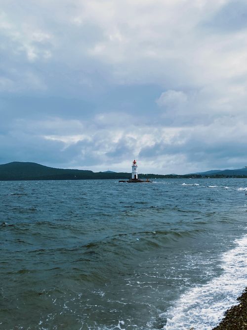 Free A picturesque lighthouse standing amidst a vast ocean under a cloudy sky, with waves gently crashing on the shore. Stock Photo