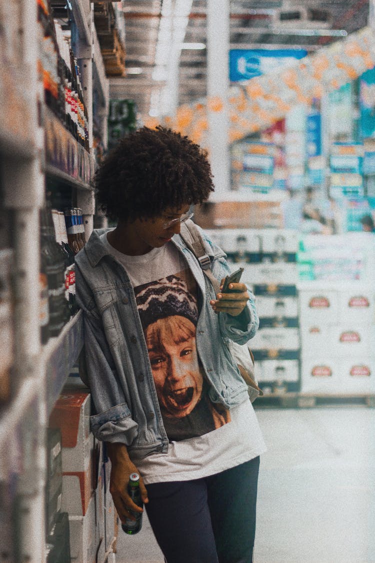 Photo Of Woman Leaning On Supermarket Shelf Carrying A Glass Bottle While Using Phone