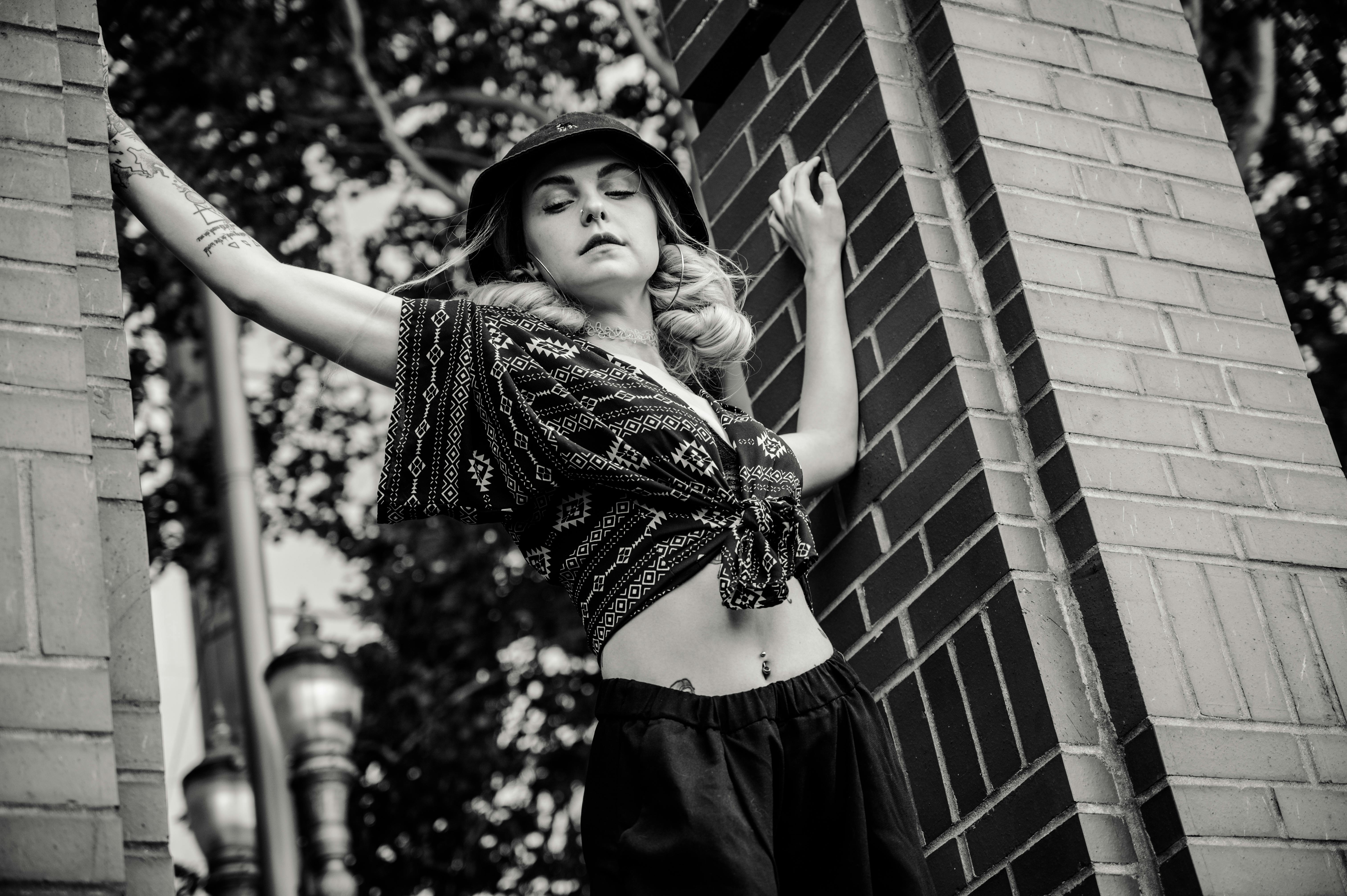 Black and white portrait of a fashionable woman posing against a brick wall outdoors.