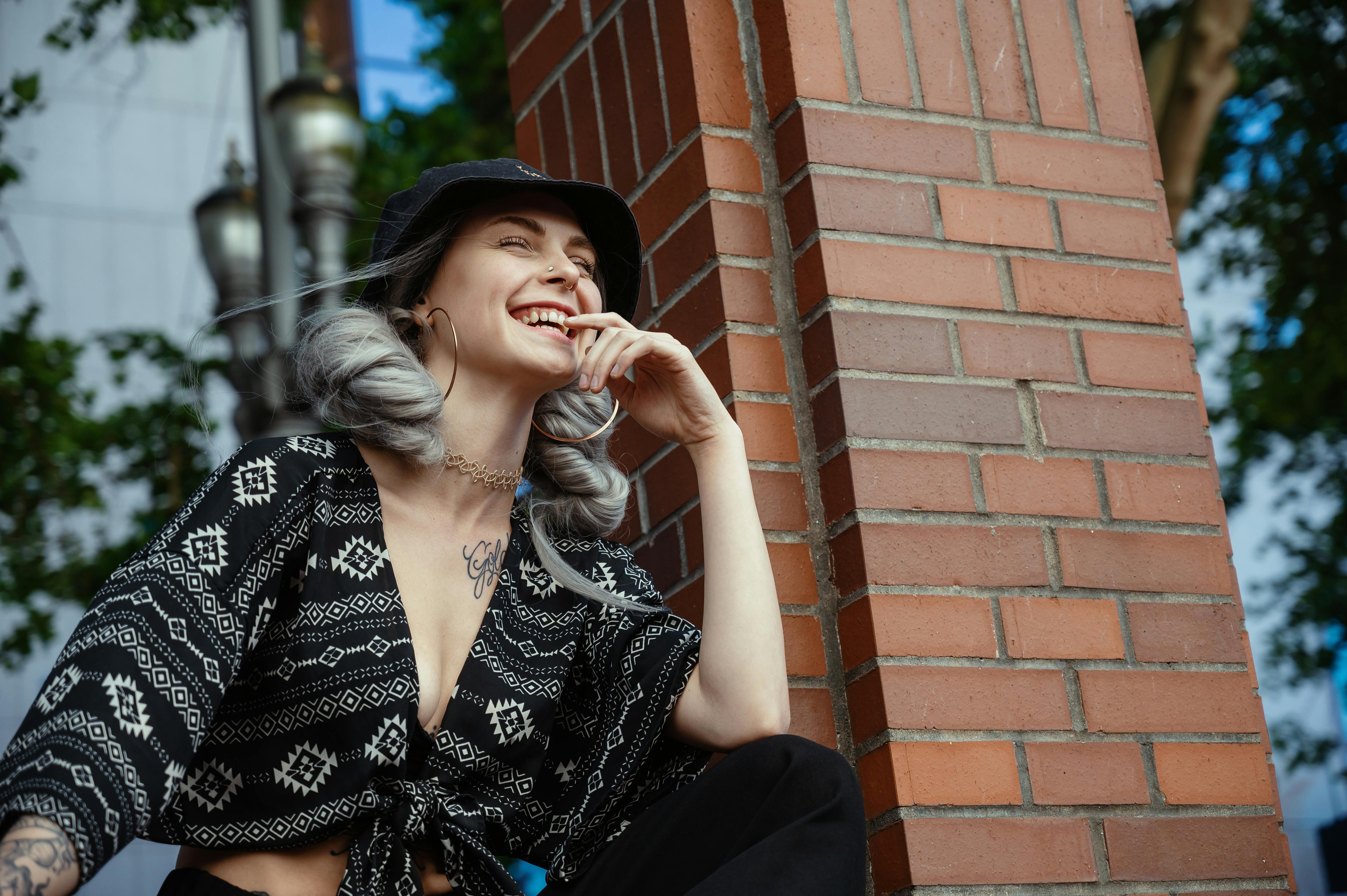 一位时尚女性身着街头服装，在阳光下快乐地倚着砖柱摆姿势，开怀大笑