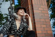 一位时尚女性身着街头服装，在阳光下快乐地倚着砖柱摆姿势，开怀大笑