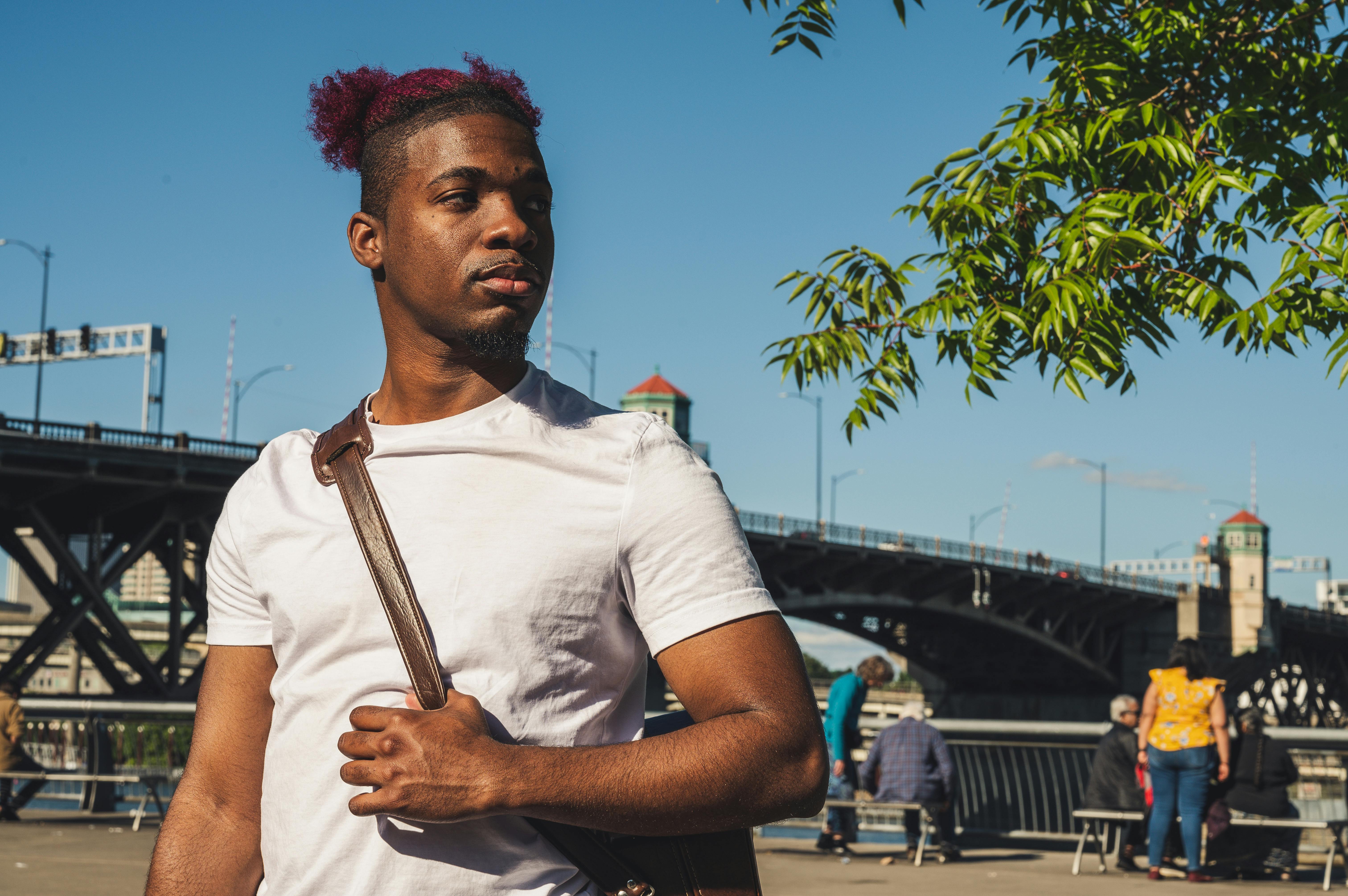 一位时尚男士斜挎着包，站在一座桥附近，背景是充满活力的城市