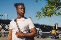 一位时尚男士斜挎着包，站在一座桥附近，背景是充满活力的城市