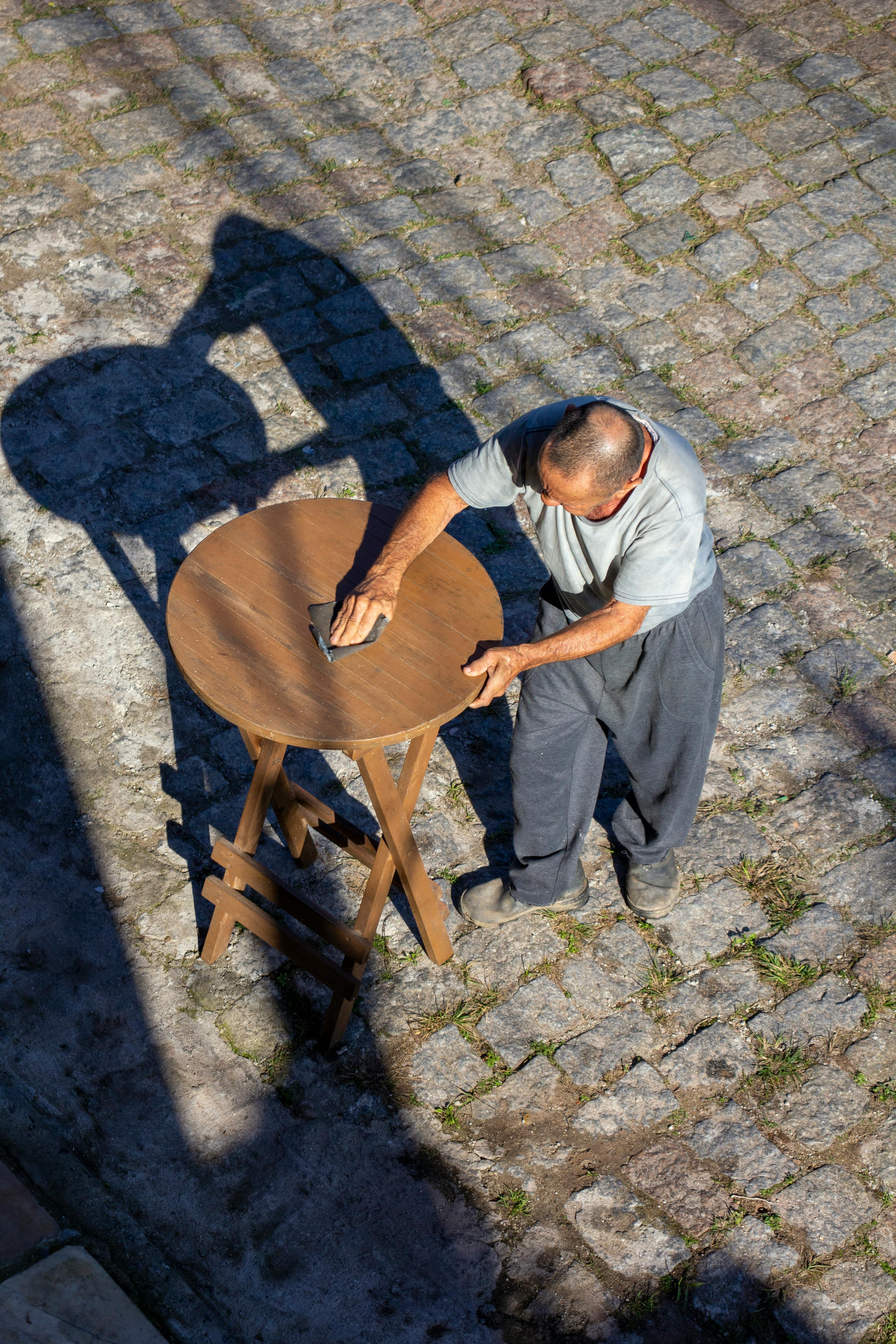 A man cleans a wooden table on a cobblestone street with strong sunlight casting shadows.