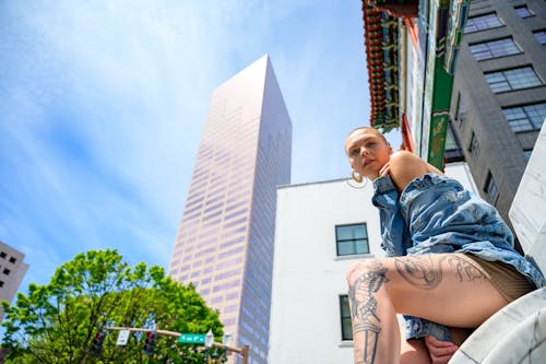Free Low Angle Photo of Woman in Blue Denim Jacket Sitting on Marble Surface Posing with Building in the Background Stock Photo