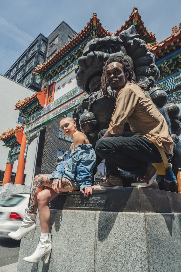 Shallow Focus Photo Of Man And Woman Sitting Beside Foo Dog Statue