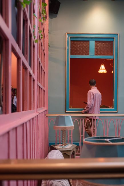 A man in a pink shirt stands in a vibrant art museum interior with pink and teal walls.
