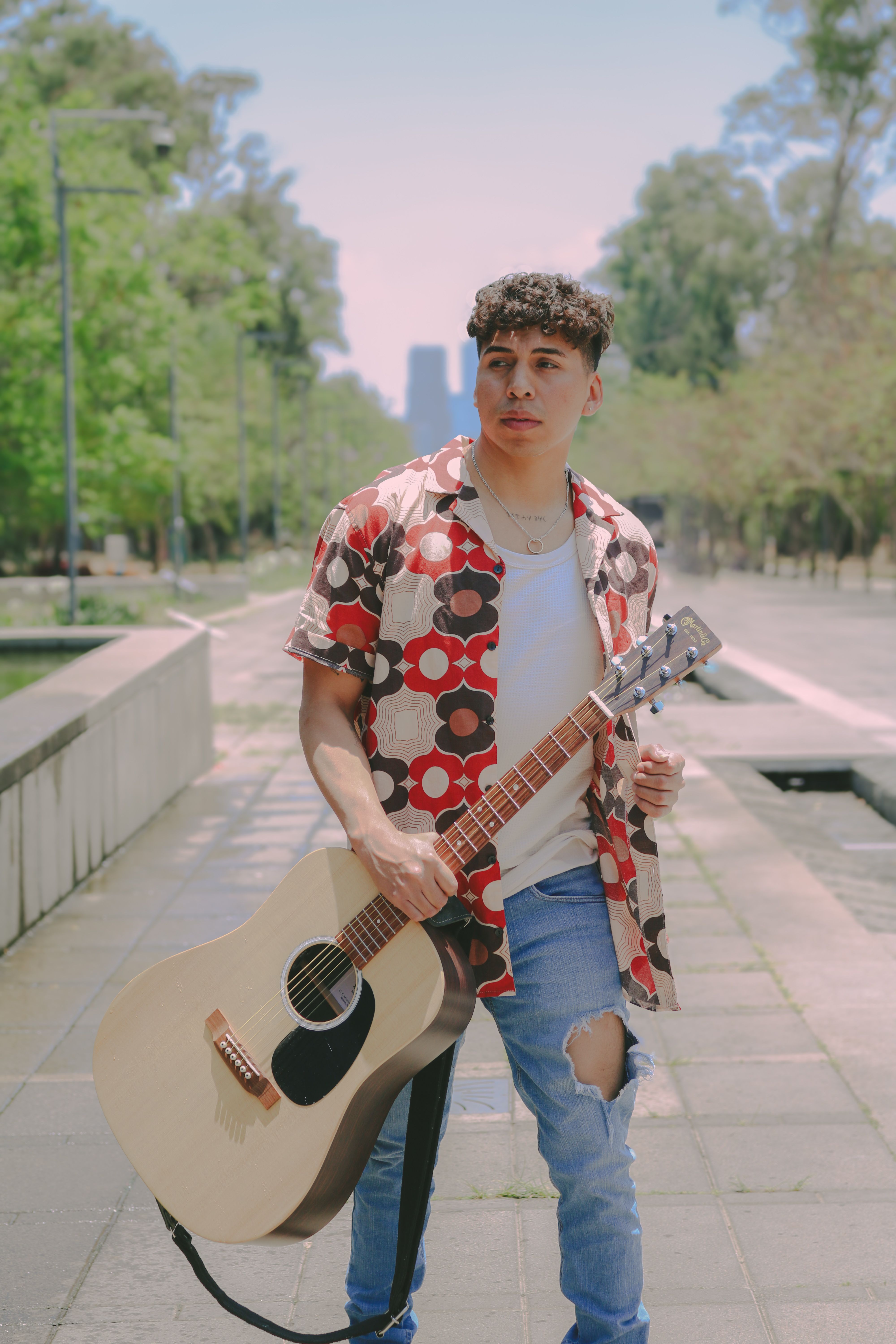 Casual young man with guitar walking down a city sidewalk on a sunny day.