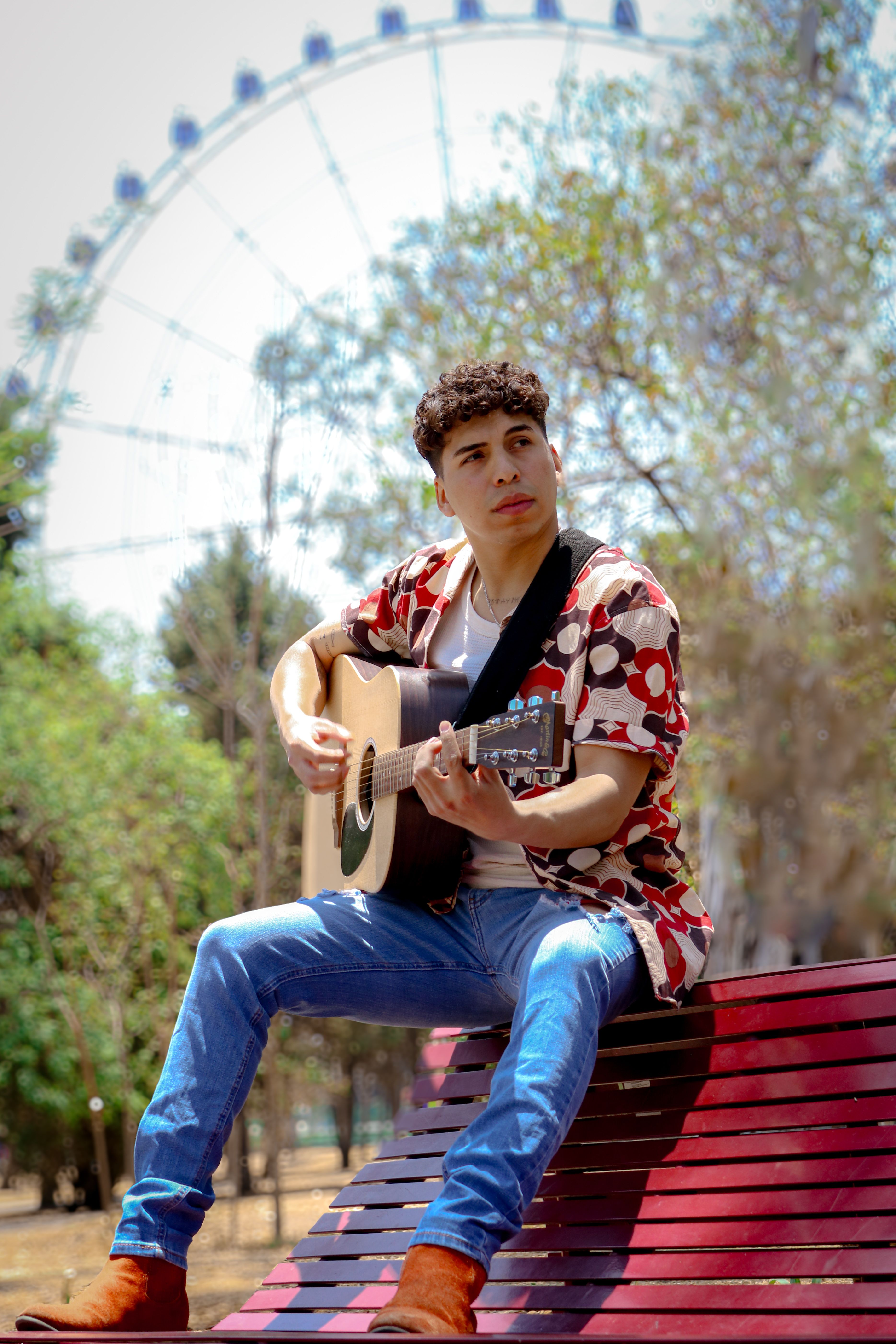 A young man sits on a red park bench playing guitar in front of a Ferris wheel.