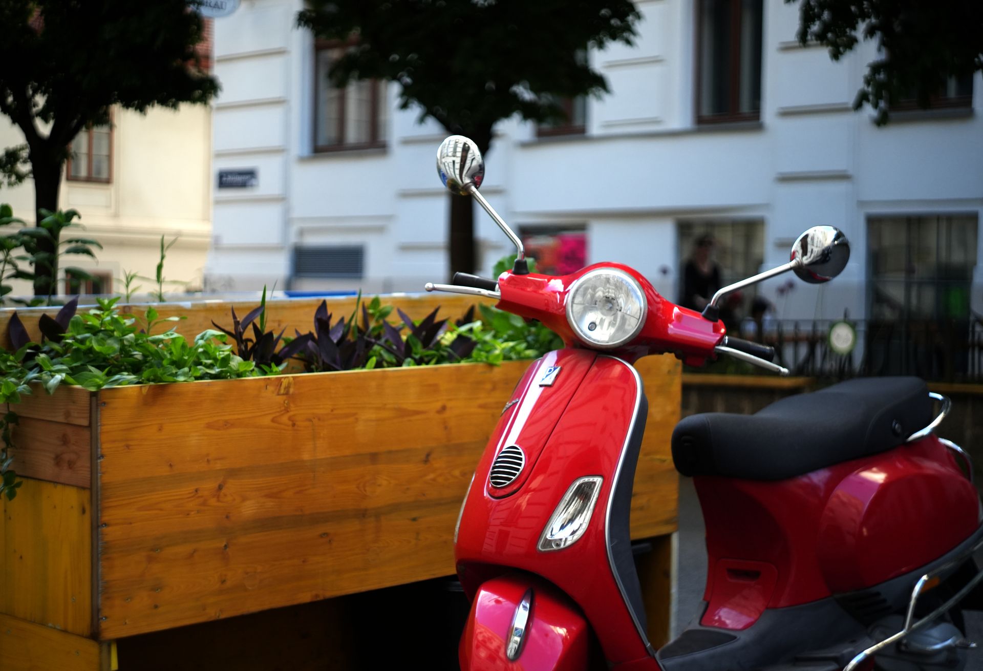 Red Vespa scooter parked in Vienna's vibrant urban streets, next to greenery and architecture.