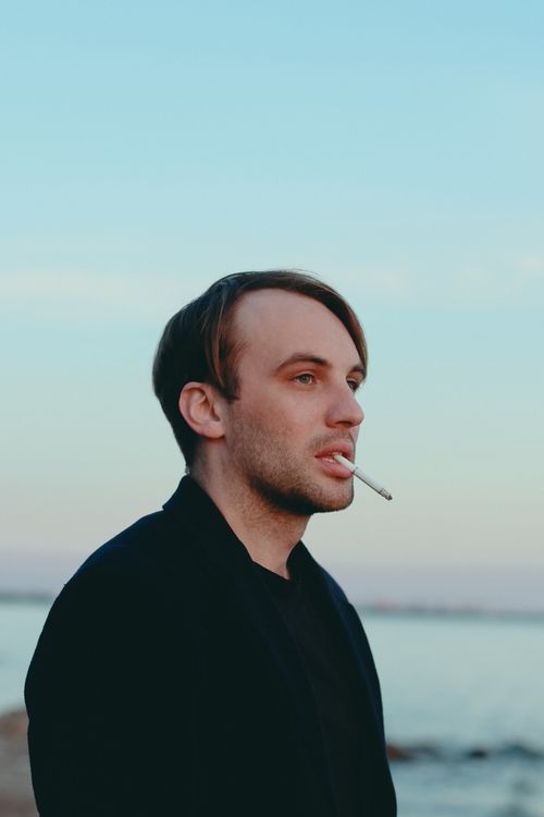 Free Portrait of a young man smoking by the sea during twilight, with a serene sky and calm waters. Stock Photo