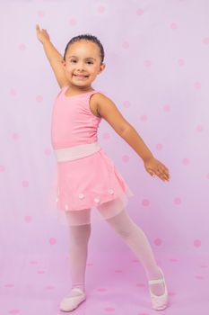 Cute young girl posing in pink ballet outfit, smiling with arms up against a polka dot background.