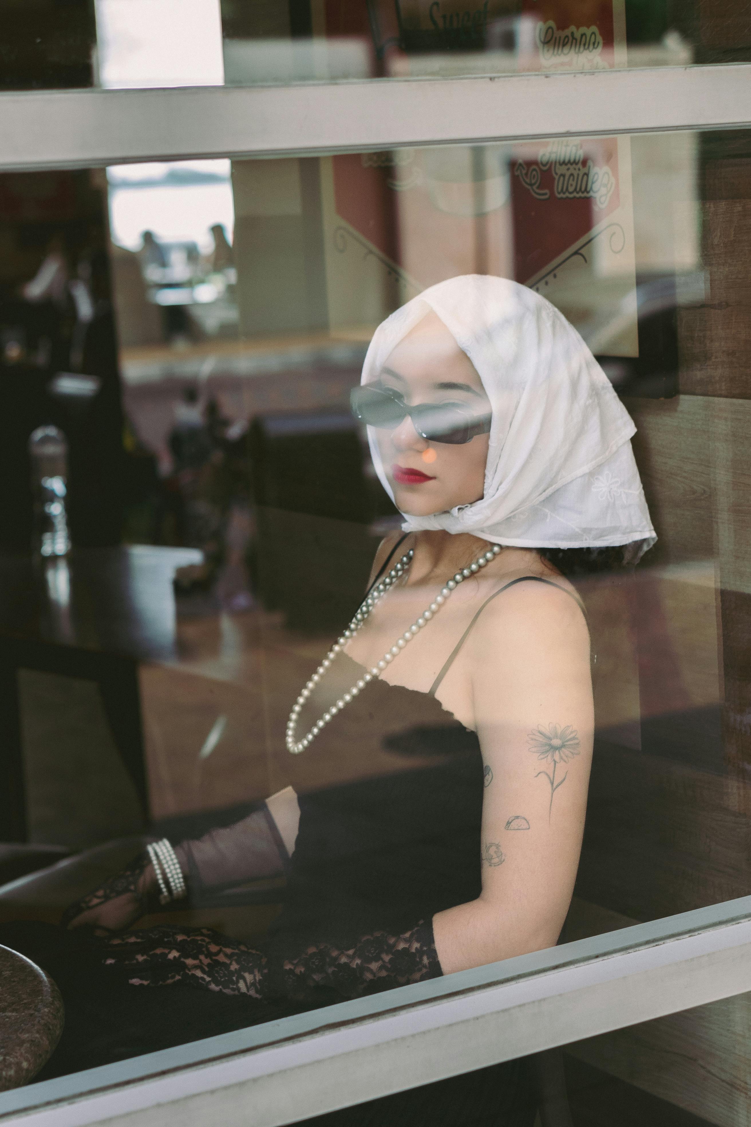 Chic woman in a black dress and headscarf gazes through window, embodying vintage elegance.