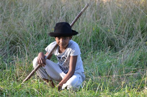 Free A young boy in a hat sits in a grassy field, holding a stick with a playful expression. Stock Photo