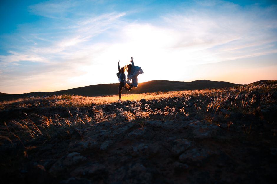 Two friends jump joyfully at sunset in a rocky field, creating a silhouette against the vibrant sky.