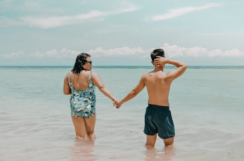 Back View Photo of Couple in Swimming Costume Holding Hand While Standing in the Water at the Beach