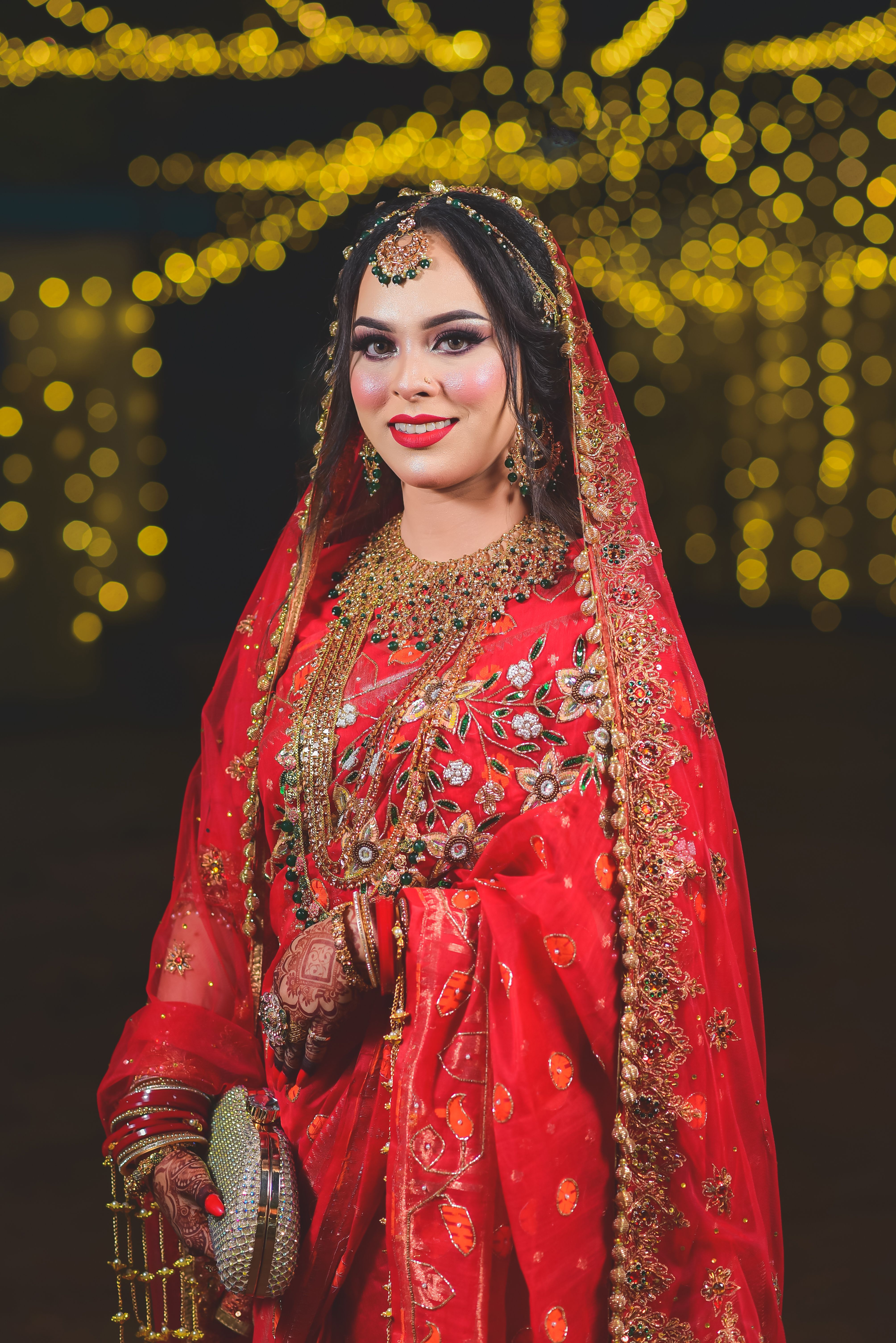 Stunning South Asian bride in vibrant red traditional attire with gold jewelry, smiling against a bokeh background.