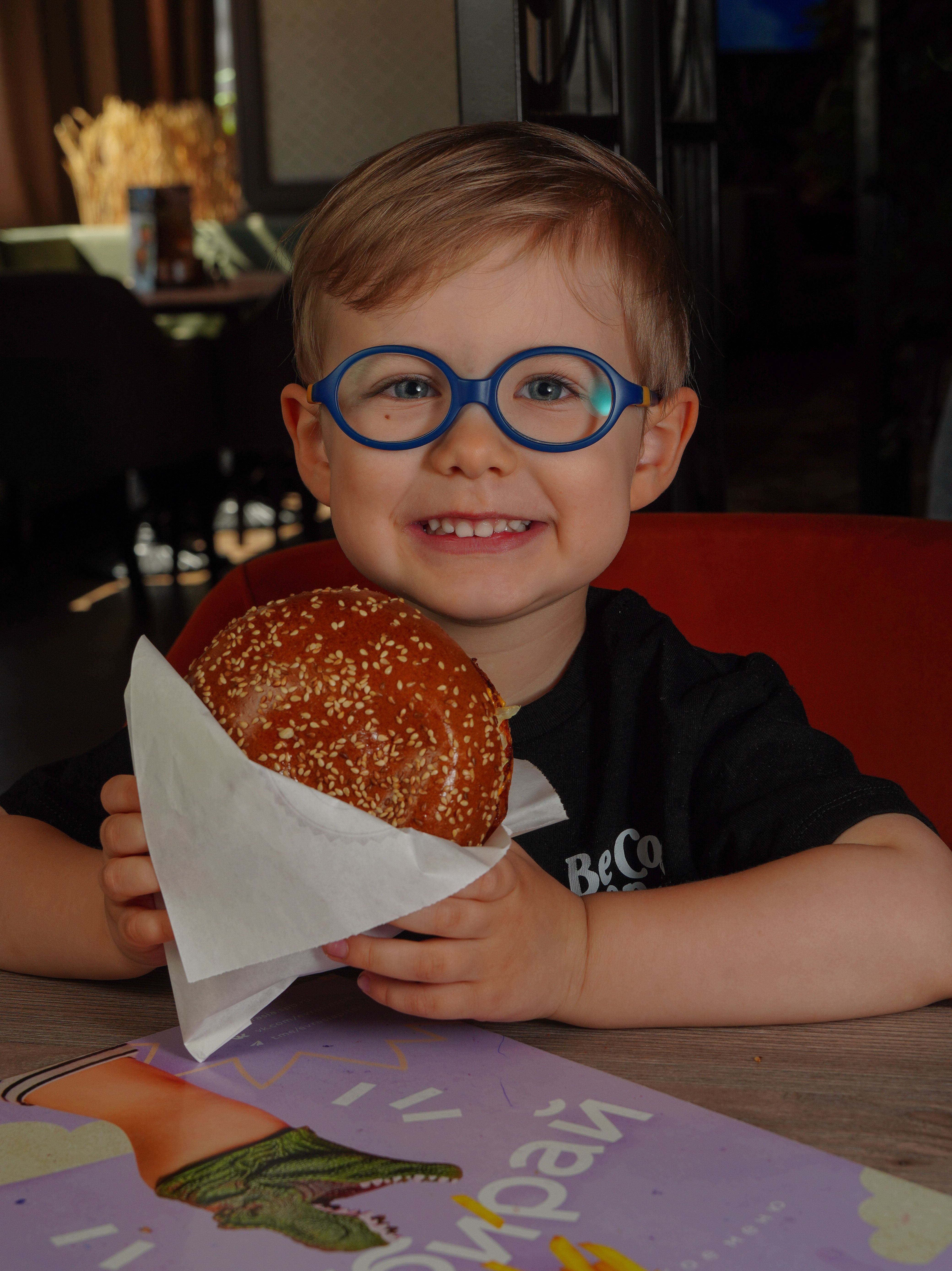 A smiling child with glasses enjoys a sesame burger inside a cozy restaurant.