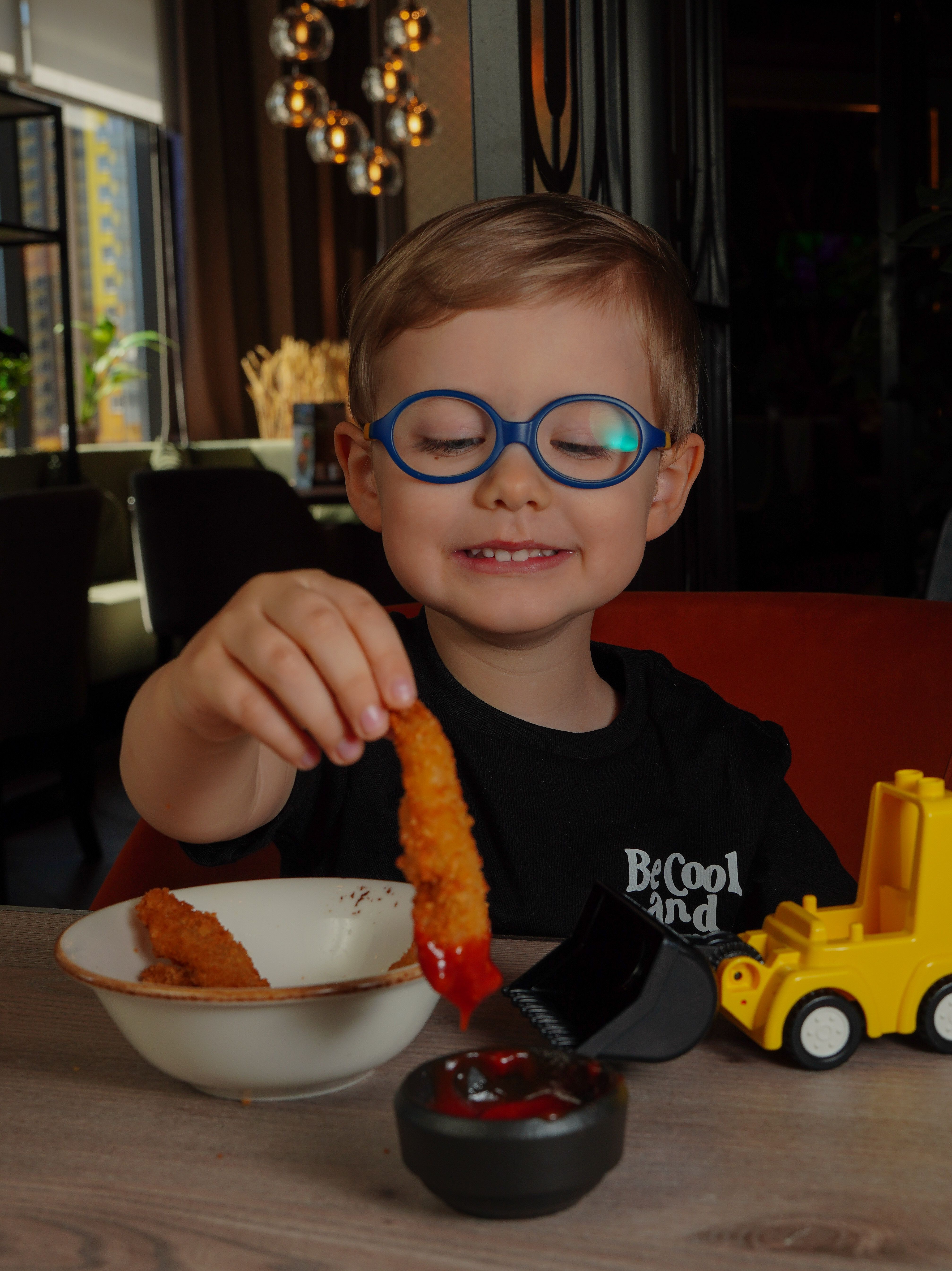 A young boy with glasses happily dips a fried chicken strip in sauce at a restaurant table.