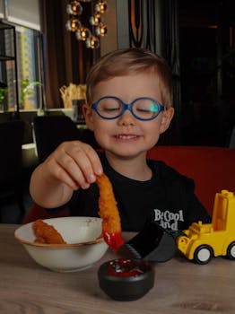 A young boy with glasses happily dips a fried chicken strip in sauce at a restaurant table.