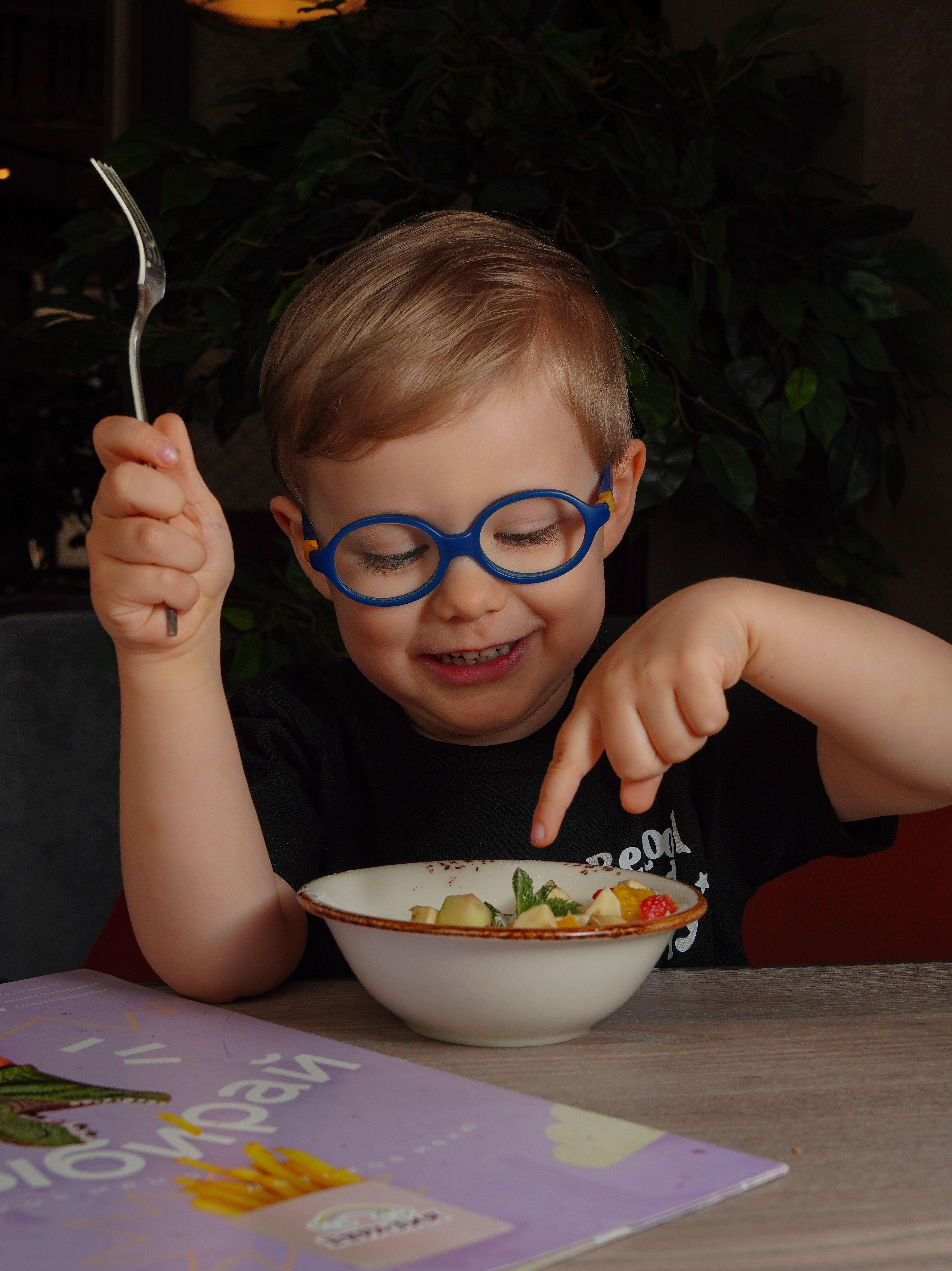 Smiling child with glasses having a healthy meal indoors, pointing and eating with joy.