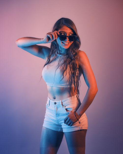 Free Fashionable woman posing confidently wearing crop top and shorts, highlighted by vibrant lighting. Stock Photo