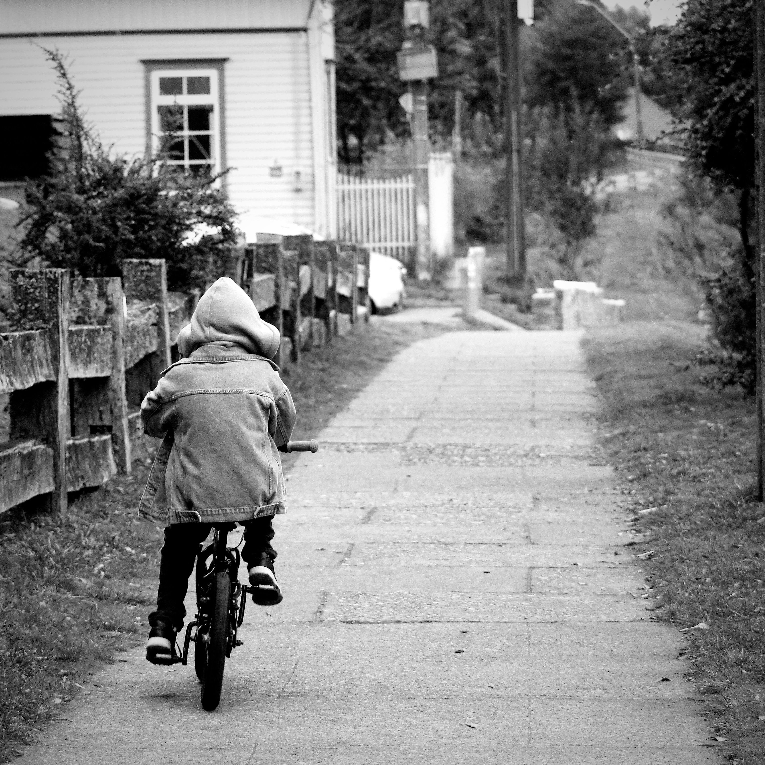 Black and white photo of a child biking on a peaceful sidewalk in a residential area.