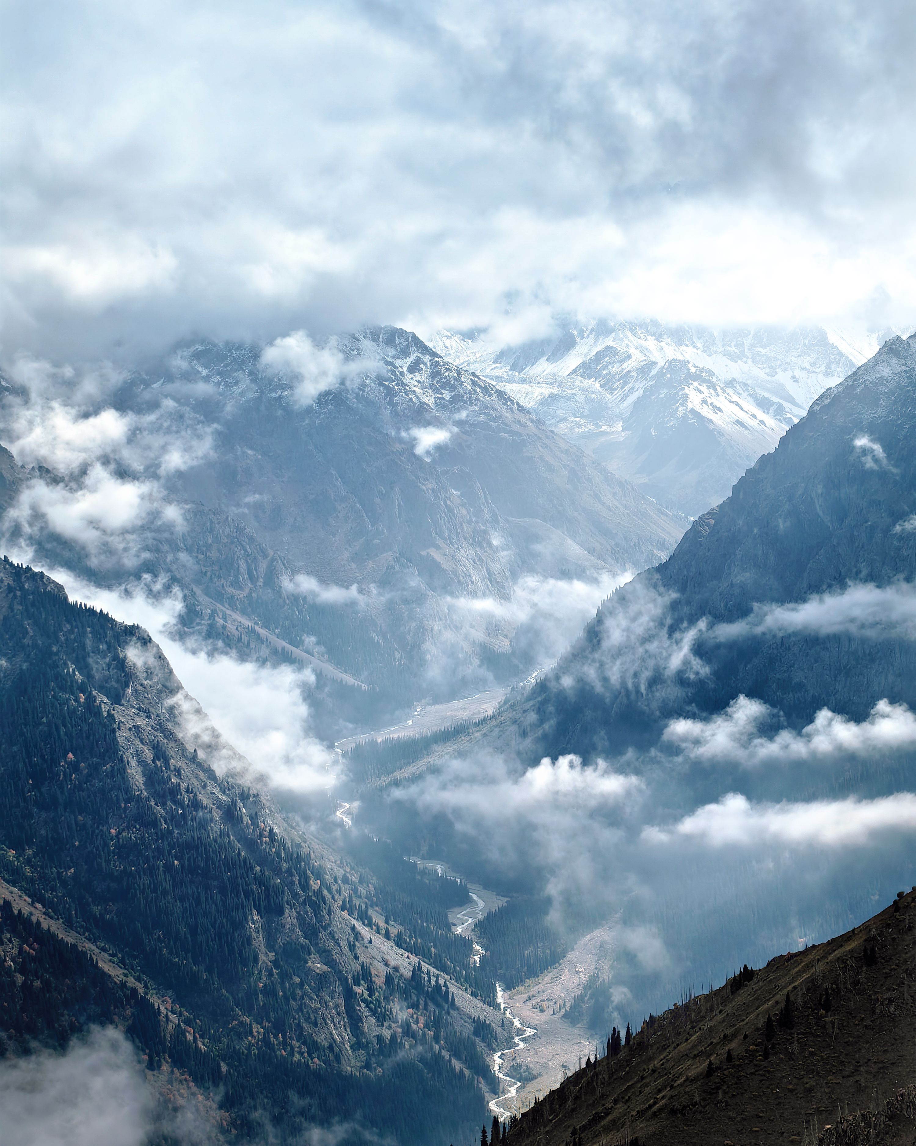 Scenic view of snow-capped mountains and valleys in Almaty, surrounded by clouds and forested slopes.