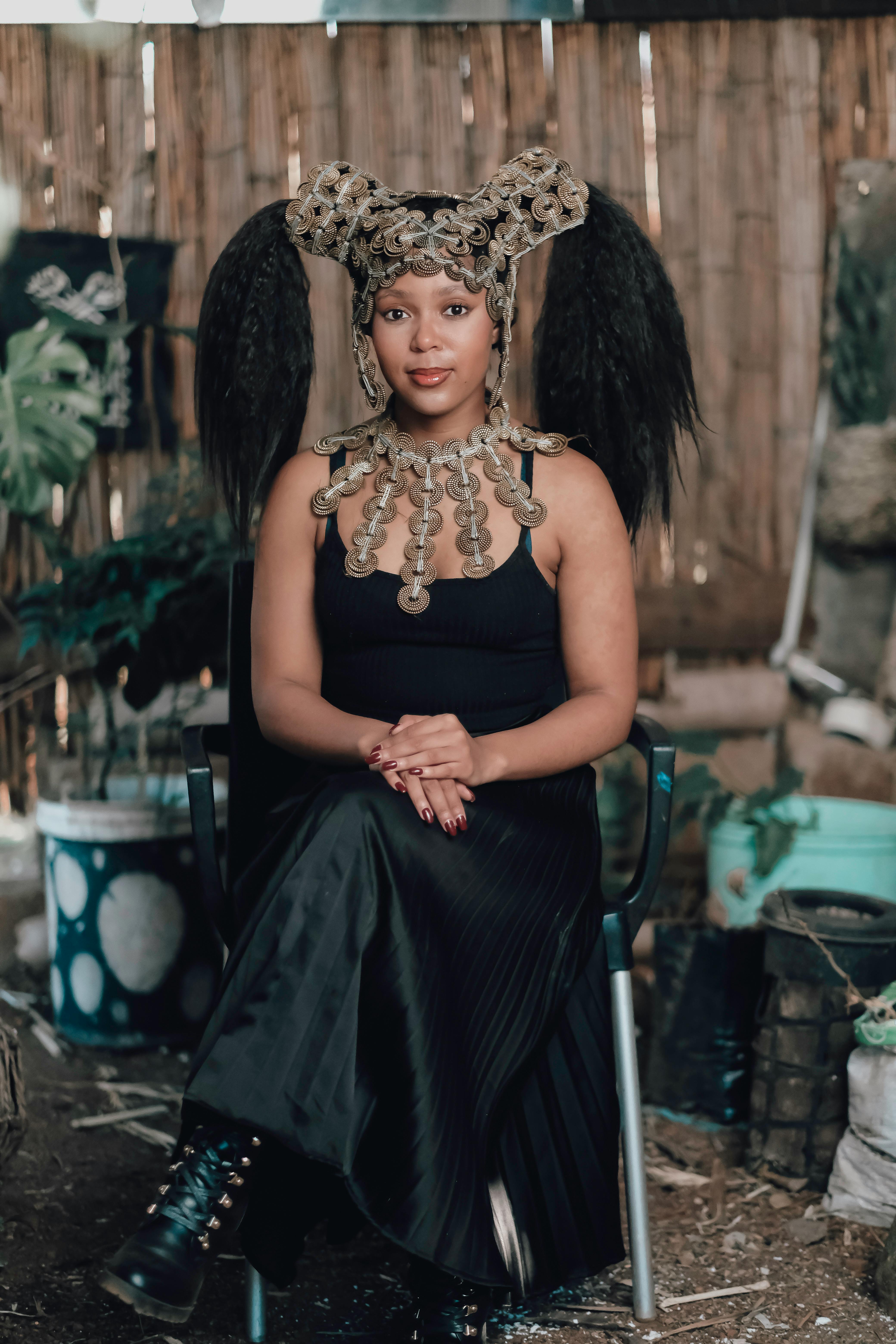 African woman in elaborate traditional headpiece and attire, seated in an artistic setting.