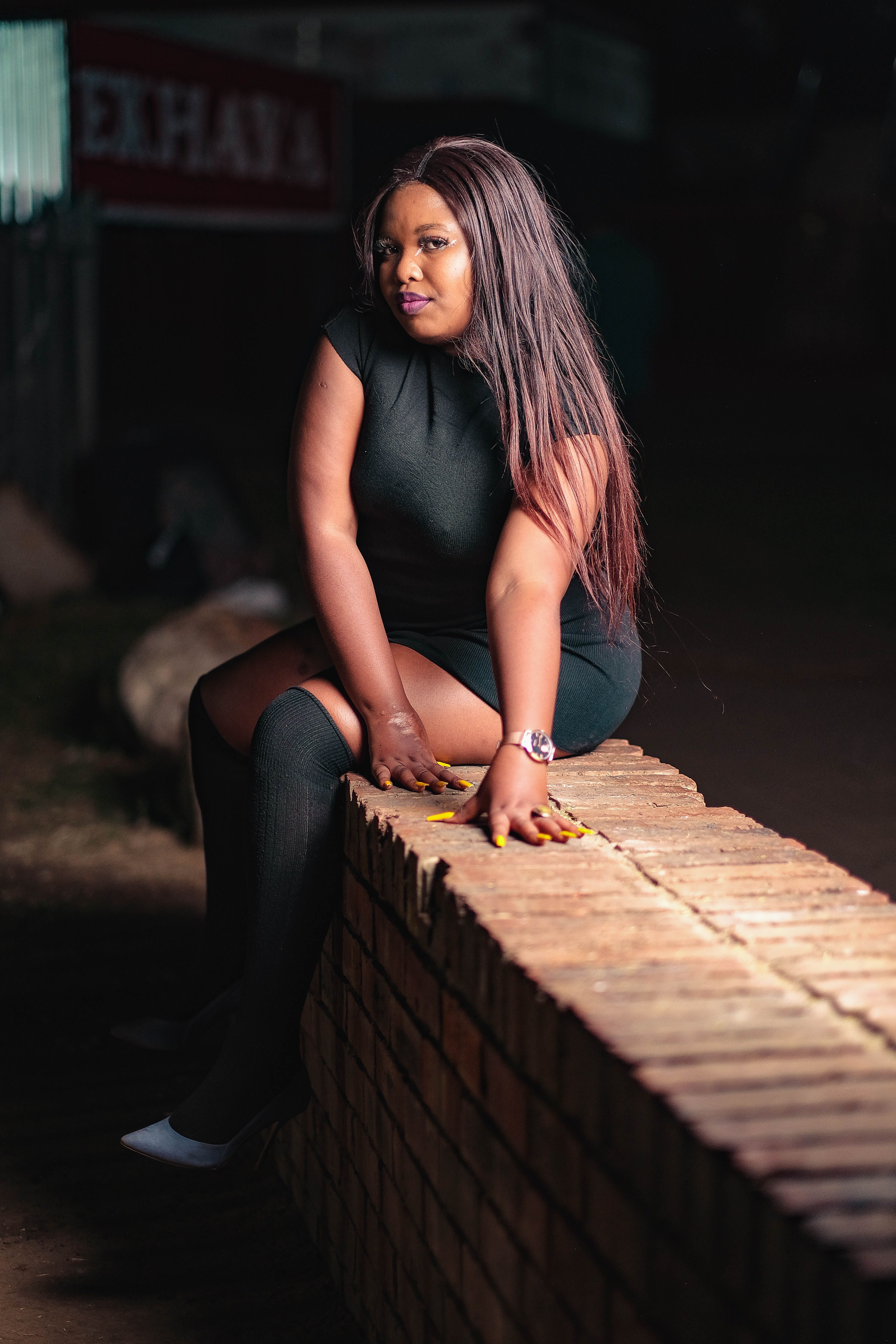 Stylish woman sitting on a brick wall at night, wearing a dark outfit.