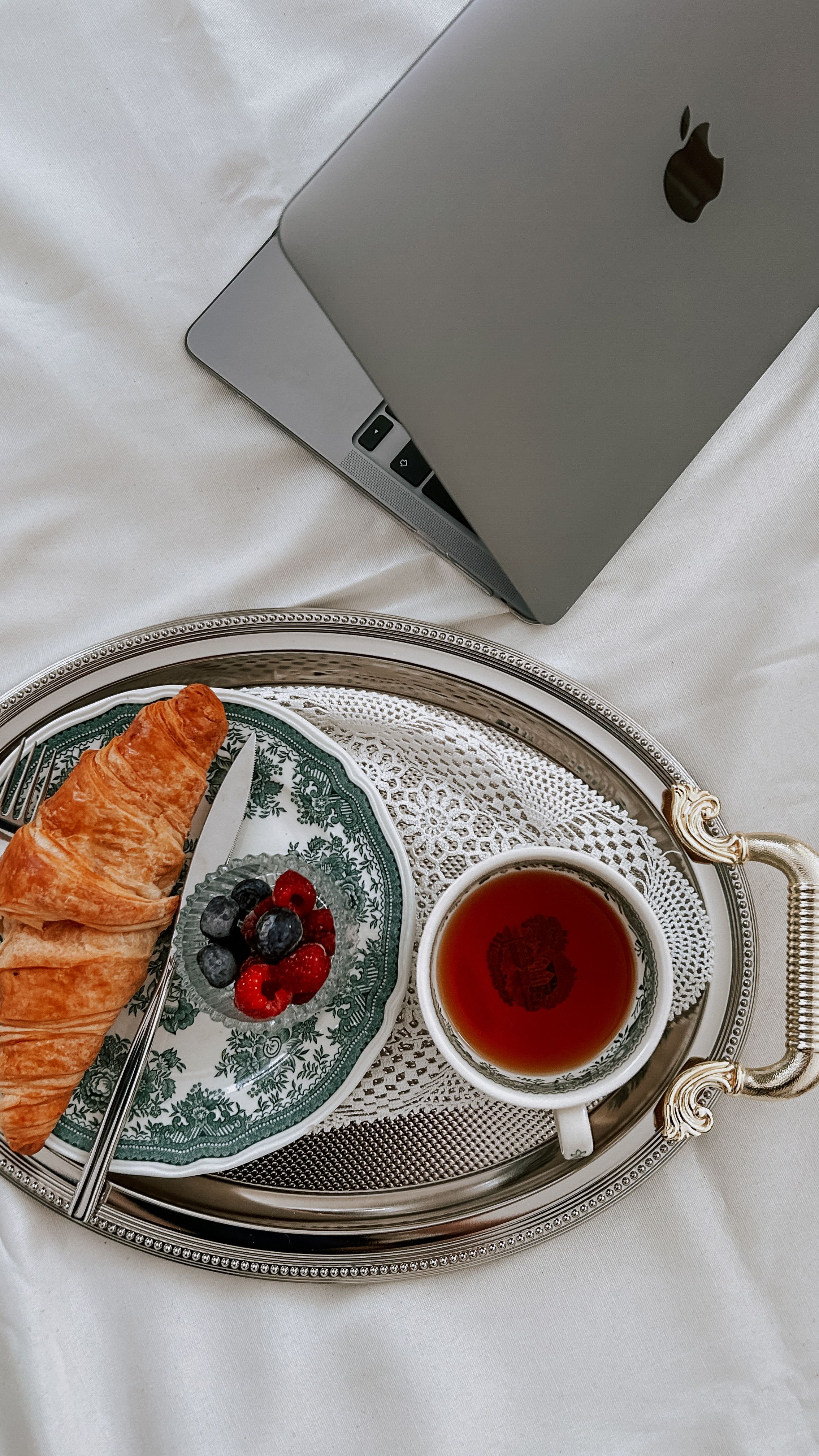 An elegant breakfast tray with croissant, berries, and tea beside a laptop on white bedding.
