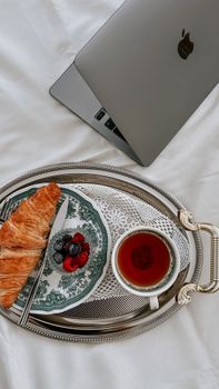 An elegant breakfast tray with croissant, berries, and tea beside a laptop on white bedding.