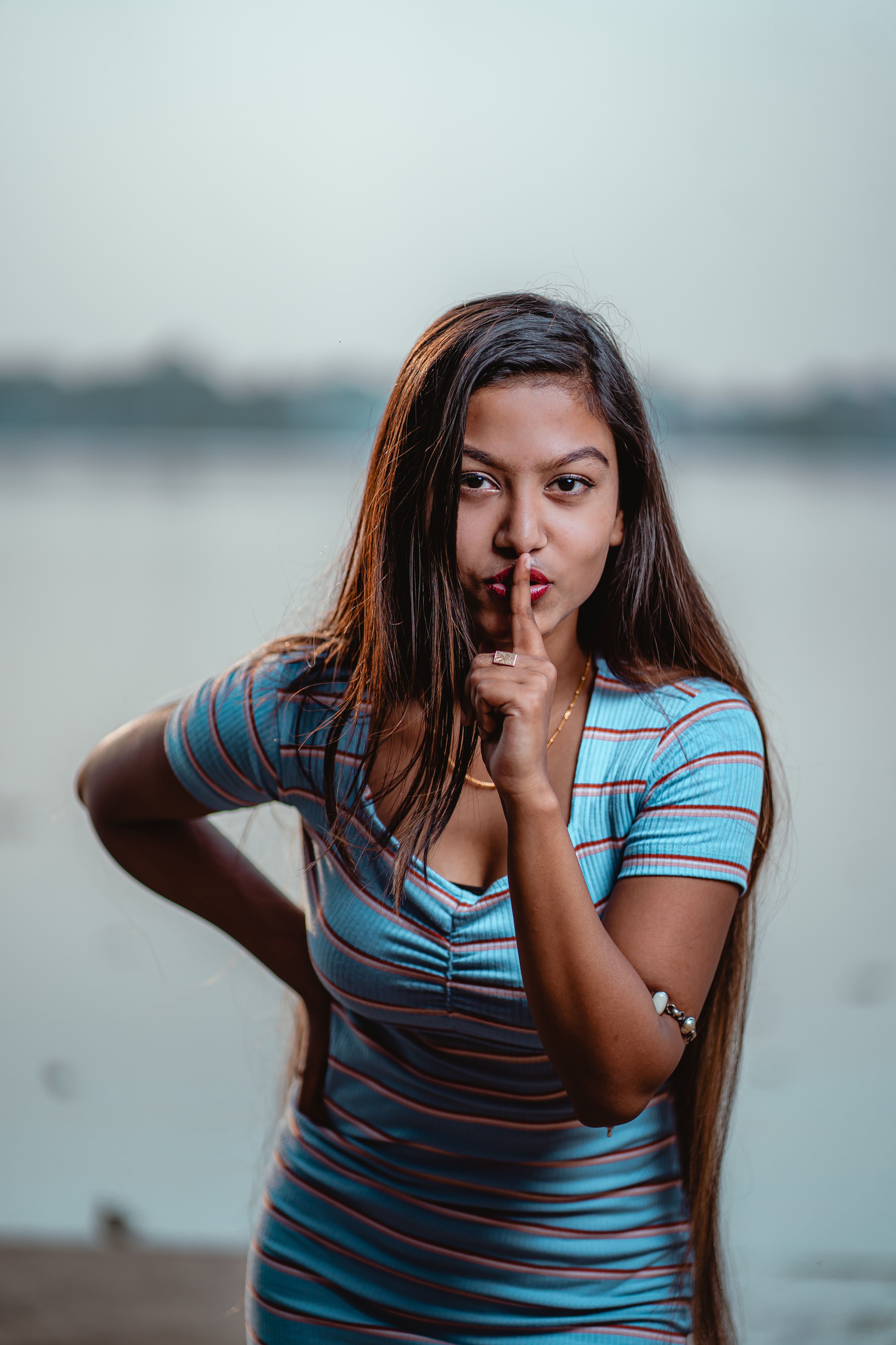 A young woman in a striped dress gestures silence with her finger to her lips near a lake.