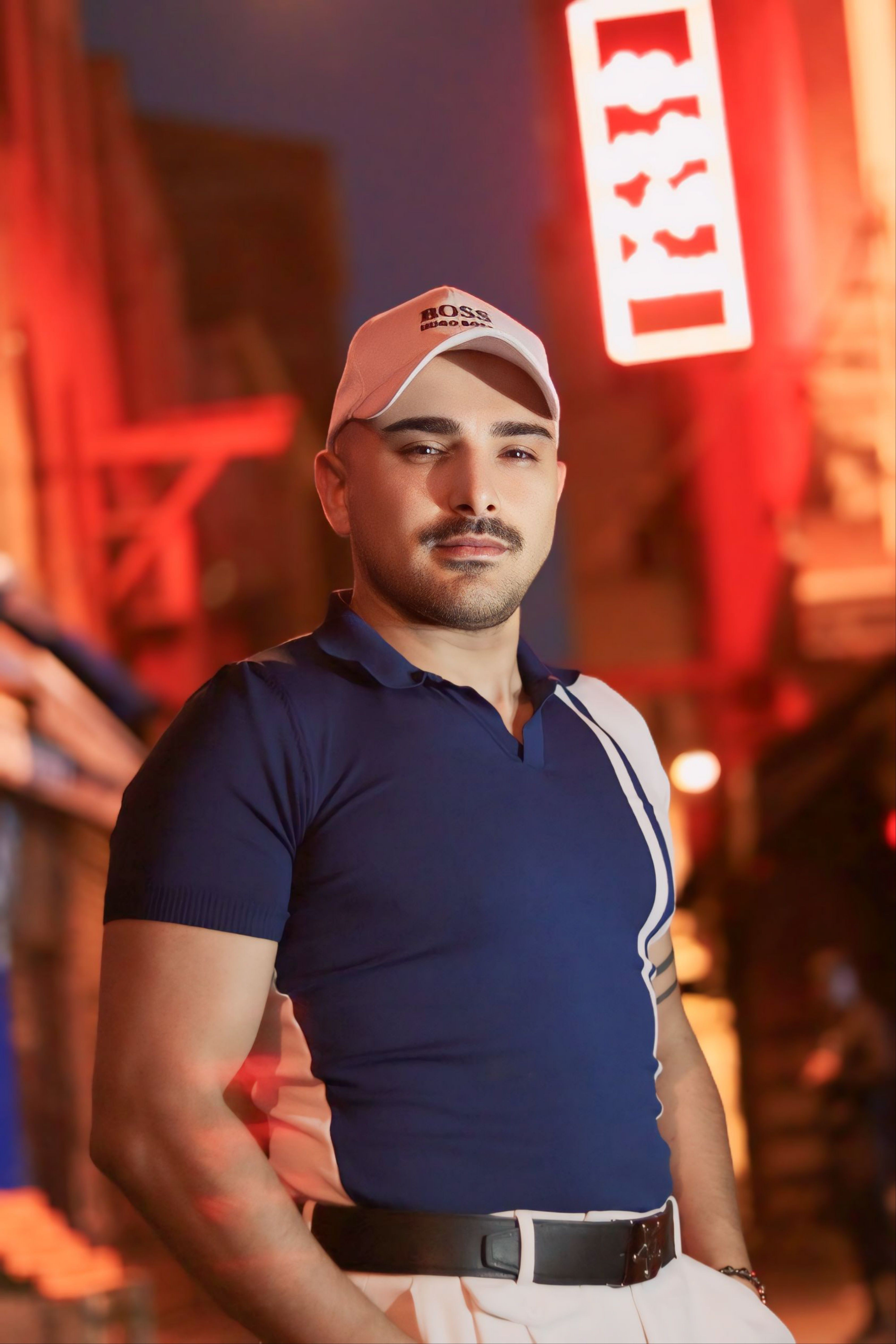 Man in blue polo shirt with white cap poses on a neon-lit city street.